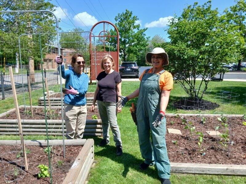 Three people stand in a community garden, smiling and holding gardening tools. Raised garden beds are visible, with plants growing.